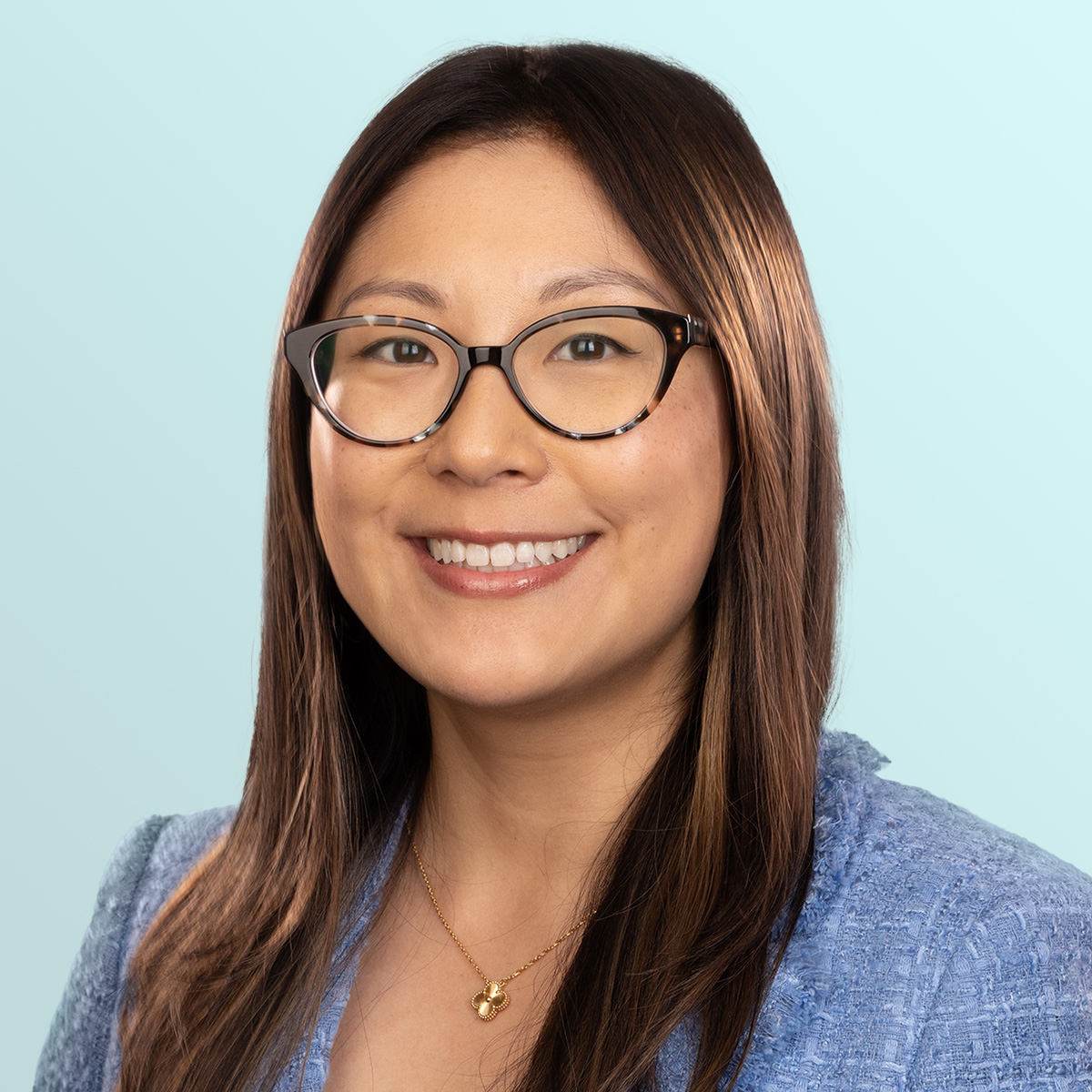 A woman with long brown hair and glasses, wearing a blue blazer and gold necklace, smiles at the camera against a light blue background.
