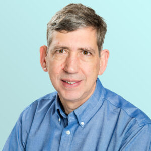 A middle-aged man with short brown hair and fair skin, wearing a blue button-up shirt, smiles gently at the camera against a light blue background.