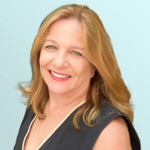 A middle-aged woman with long, light brown hair smiles warmly at the camera. She is wearing a sleeveless black top with beige trim, set against a light blue background.