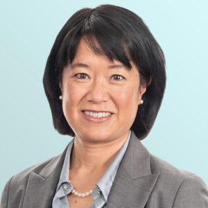 Portrait of an Asian woman with short black hair, wearing a gray blazer, light blue shirt, pearl necklace, and small pearl earrings, smiling in front of a light blue background.