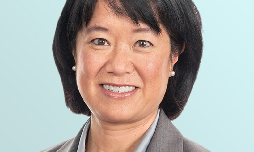 Portrait of an Asian woman with short black hair, wearing a gray blazer, light blue shirt, pearl necklace, and small pearl earrings, smiling in front of a light blue background.