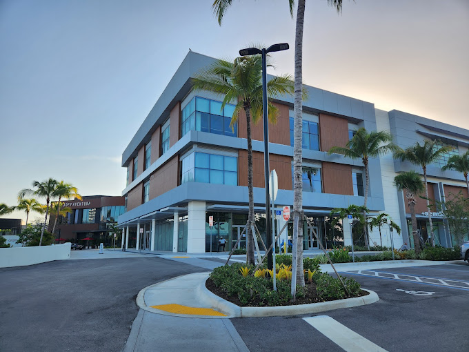A modern, multi-story building with large windows and palm trees in front; the sun is setting, casting a warm light, and there are empty parking spaces and a curved driveway in the foreground.