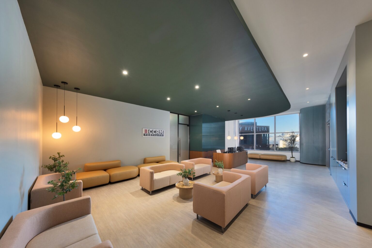 A modern waiting room with beige armchairs, round tables with plants, pendant lights, and a sleek reception desk near large windows letting in natural light.