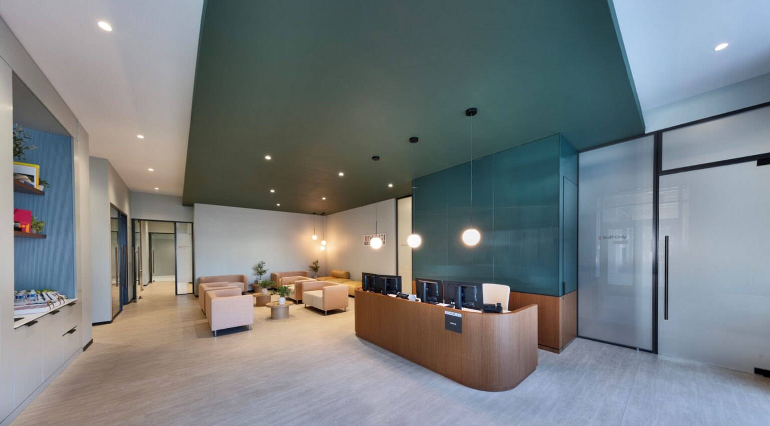 Modern office reception area with a curved wooden desk, computer monitors, and pendant lights. Beige armchairs and small tables are arranged in a waiting area, with plants and contemporary decor under a dark green ceiling.