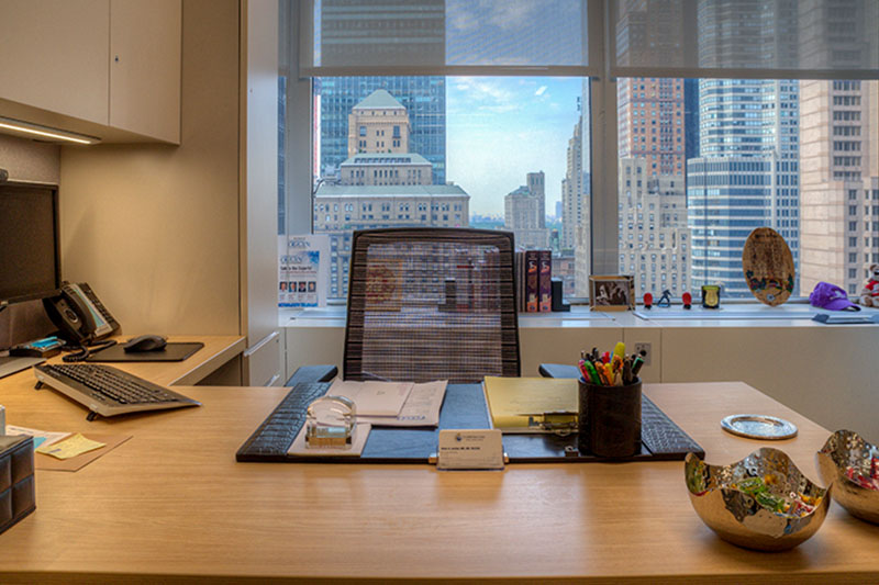 A modern office desk with a mesh chair, computer, phone, office supplies, and papers, set beside a large window overlooking tall city buildings in daylight.