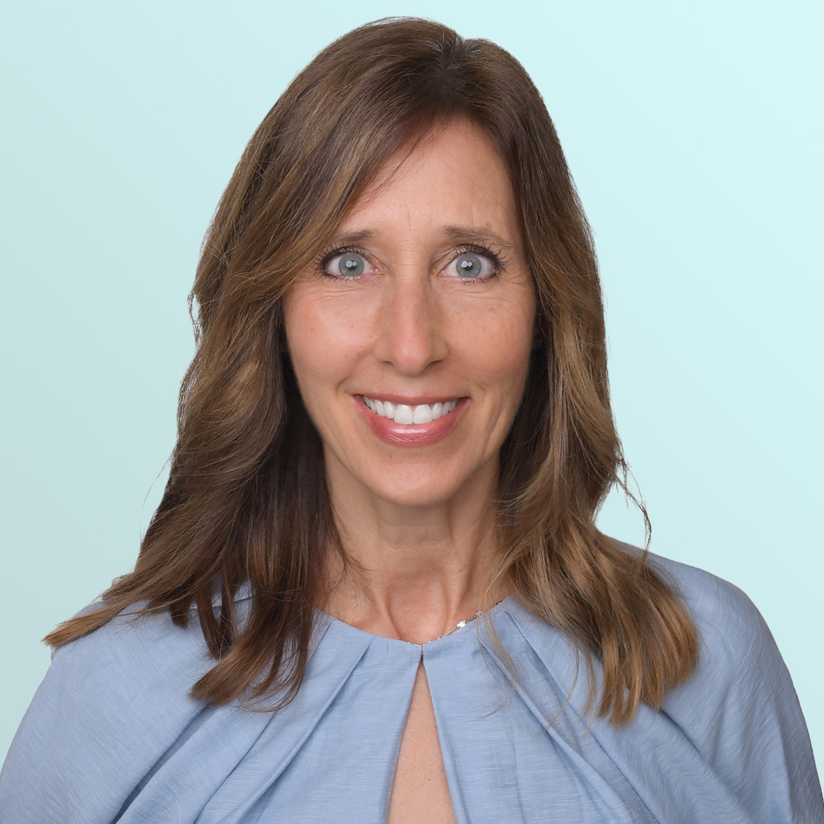 A woman with long brown hair and blue eyes smiles at the camera. She is wearing a light blue top and is posed against a pale blue background.