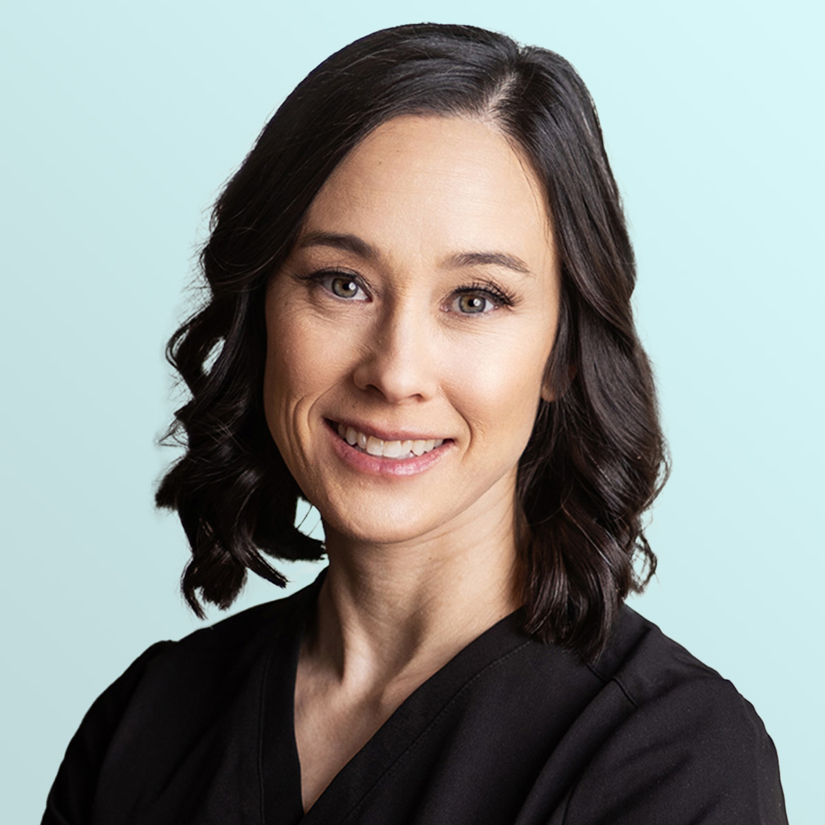 A woman with shoulder-length dark brown wavy hair, wearing a black top, smiles at the camera against a light blue background.