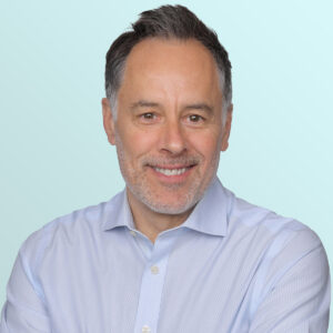 Smiling man with short dark hair and light stubble, wearing a light blue button-up shirt, standing against a pale blue gradient background.