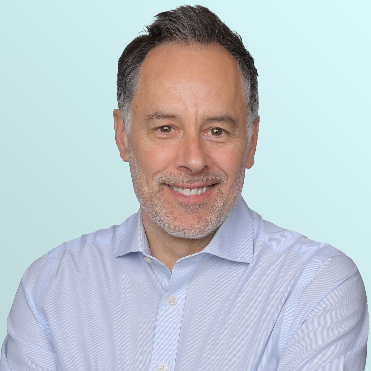 Smiling man with short dark hair and light stubble, wearing a light blue button-up shirt, standing against a pale blue gradient background.