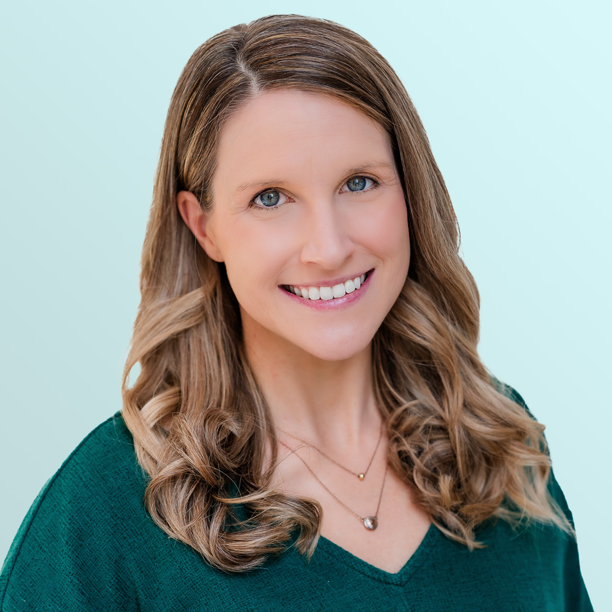 A woman with long, wavy light brown hair wearing a green top and two delicate necklaces smiles at the camera against a light blue background.