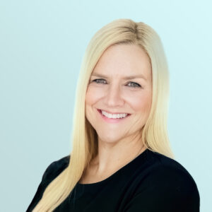 A woman with long straight blonde hair, wearing a black top, smiles confidently at the camera against a light blue background.