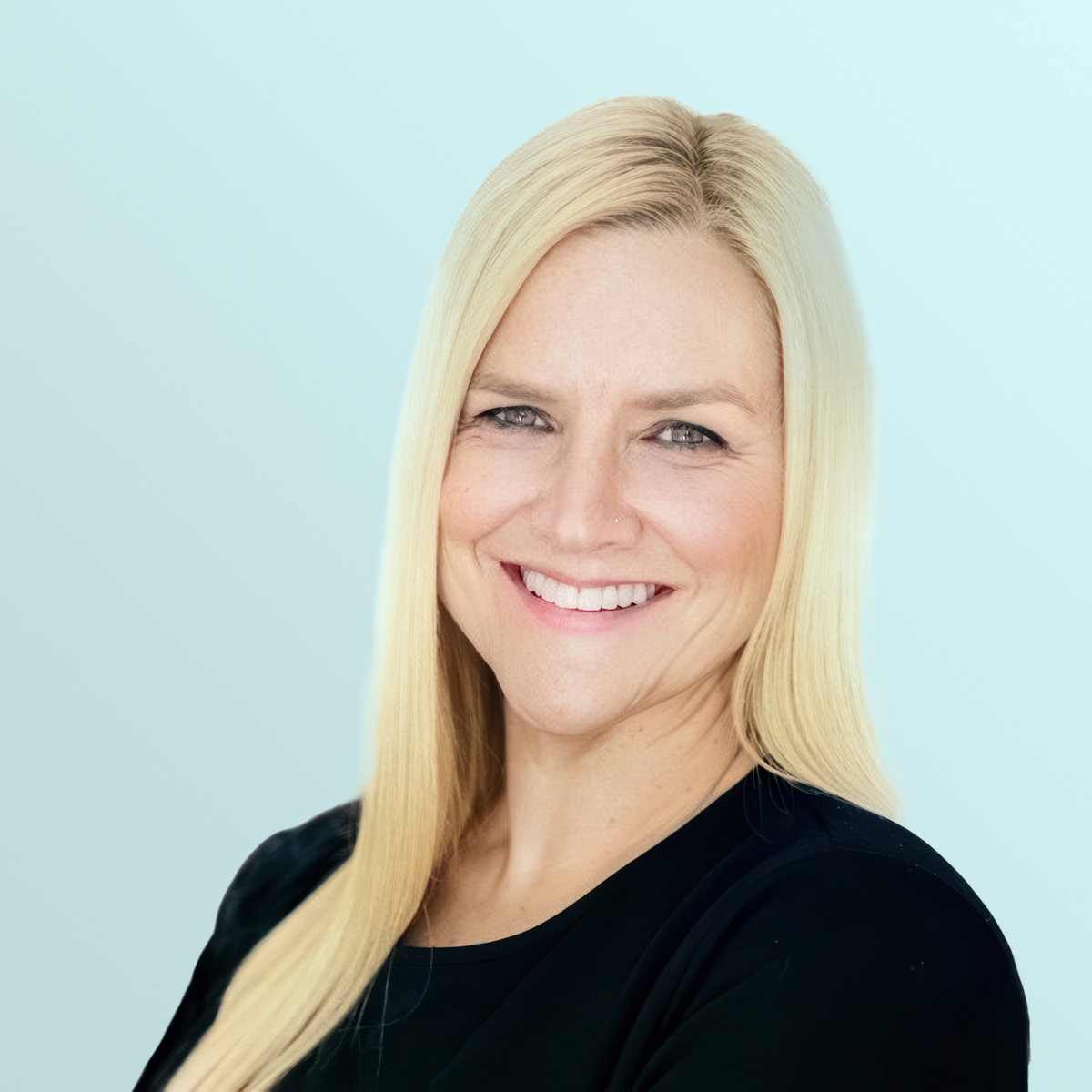 A woman with long straight blonde hair, wearing a black top, smiles confidently at the camera against a light blue background.