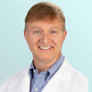 A smiling man with light brown hair wearing a white lab coat and a light blue collared shirt, posed in front of a light blue gradient background.