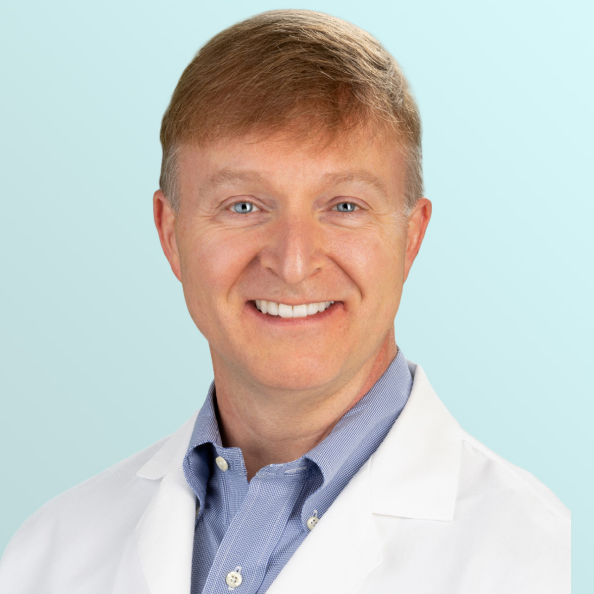 A smiling man with light brown hair wearing a white lab coat and a light blue collared shirt, posed in front of a light blue gradient background.