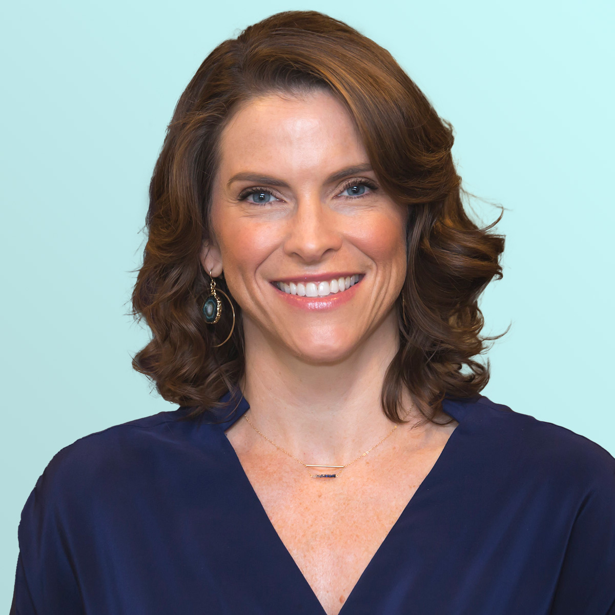 A woman with wavy brown hair and blue eyes wears a navy blouse and a pendant necklace, smiling in front of a light blue background.