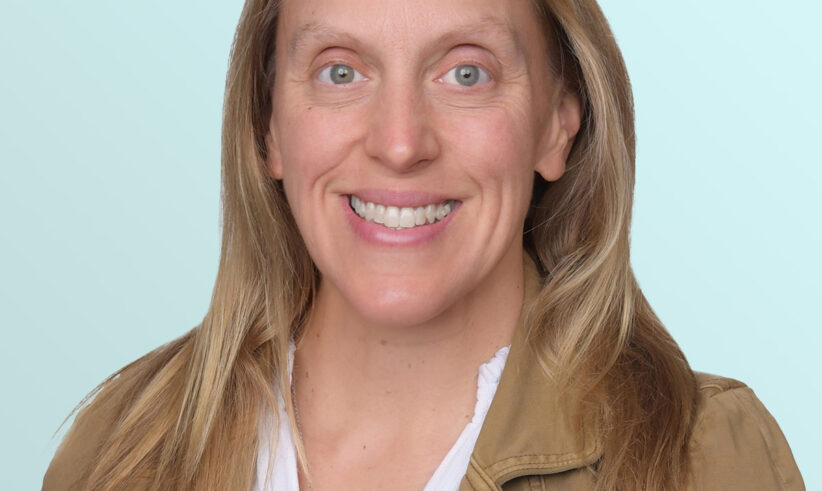 A woman with long light brown hair, wearing a tan jacket over a white blouse, smiles at the camera against a light blue background.