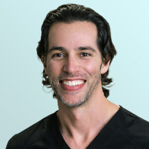 A man with dark wavy hair and light stubble smiles at the camera. He is wearing a black V-neck shirt and is posed in front of a light blue background.