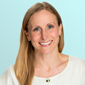 A smiling woman with long blonde hair wearing a white top and pearl earrings, posing in front of a light blue background.