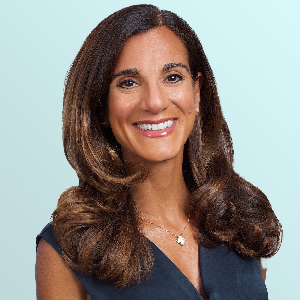 A woman with long, wavy brown hair and a wide smile wears a navy blue sleeveless top, a delicate flower pendant necklace, and pearl earrings, posing against a light blue background.