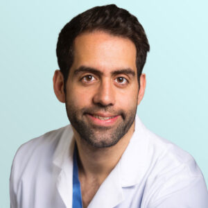 A man with short dark hair and a beard, wearing a white medical coat, smiles at the camera against a light blue background.