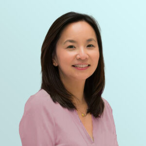 A woman with straight dark hair and a light pink blouse smiles at the camera against a pale blue background.