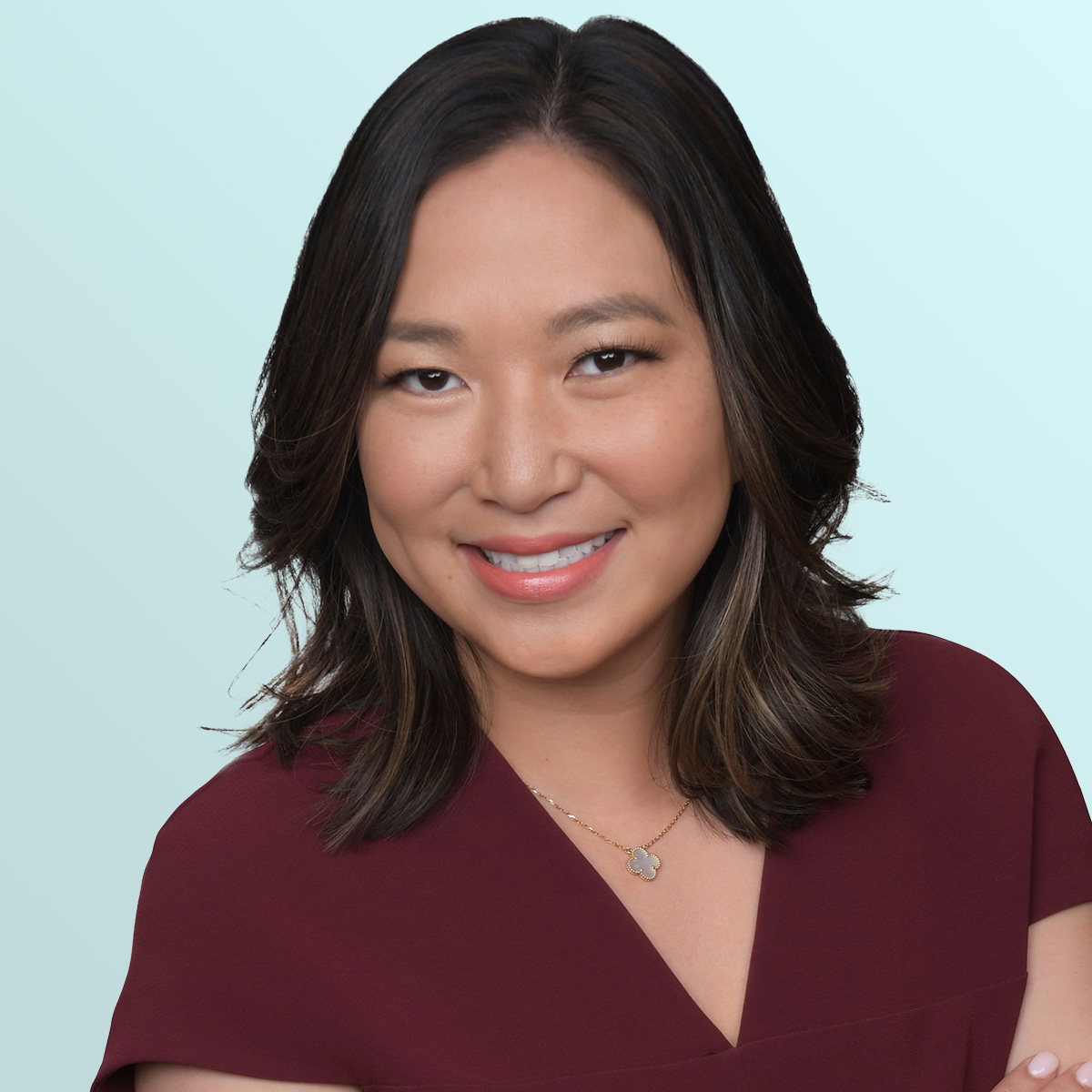 A woman with medium-length dark hair smiles at the camera. She is wearing a maroon top and a delicate necklace with a pendant. The background is a light blue gradient.