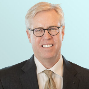 A middle-aged man with blond hair and glasses is smiling. He is wearing a dark suit, white shirt, and a tan patterned tie, with a light blue background behind him.