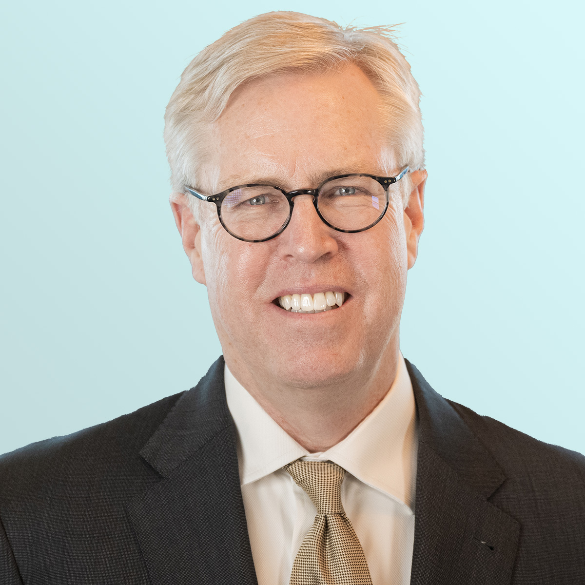 A middle-aged man with blond hair and glasses is smiling. He is wearing a dark suit, white shirt, and a tan patterned tie, with a light blue background behind him.