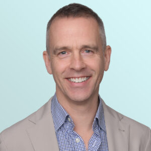 A smiling man with short hair wearing a light beige blazer over a blue and white checkered shirt, standing in front of a plain light blue background.