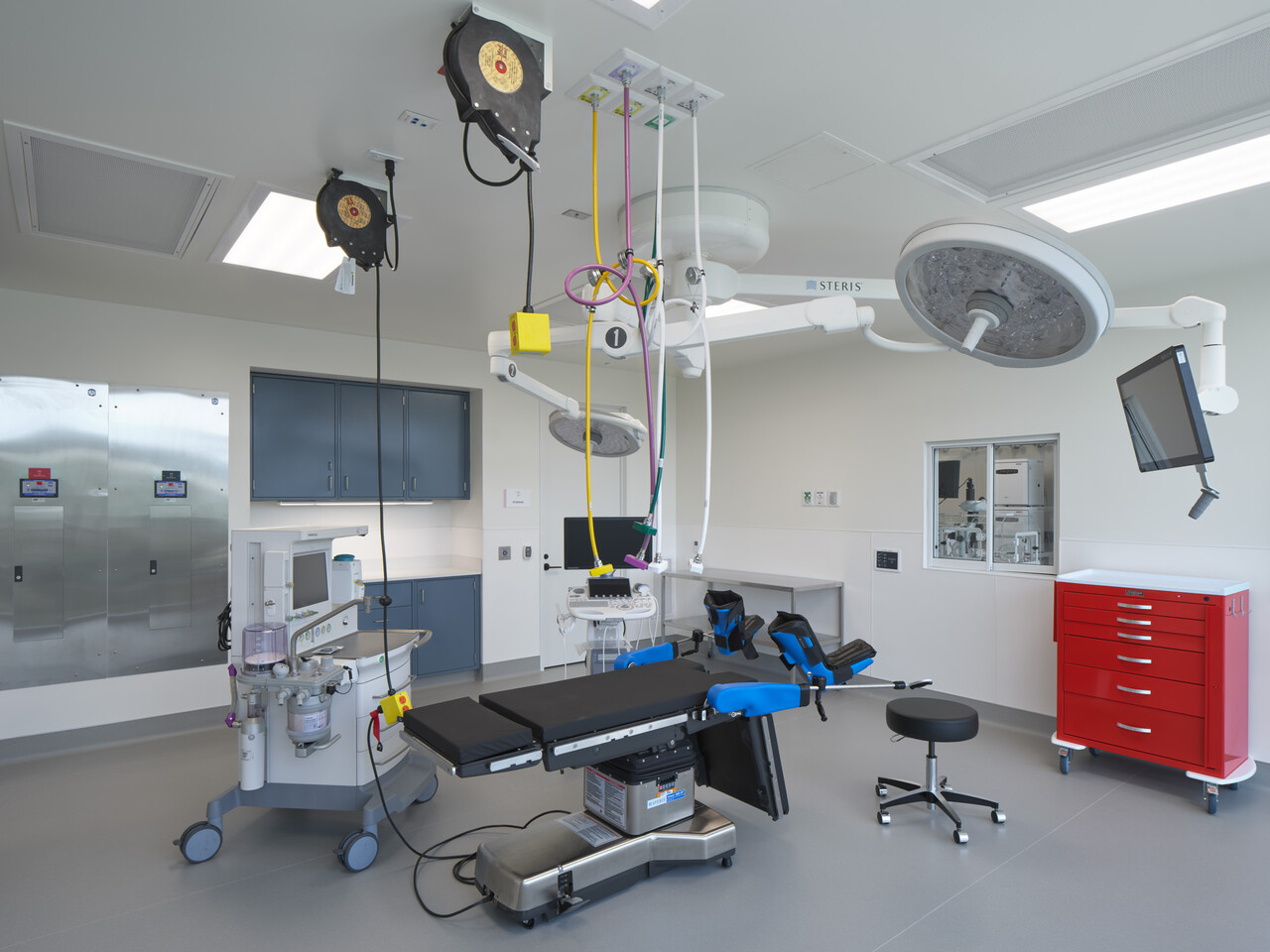 Modern operating room with an empty surgical table, overhead lights, medical equipment, monitors, red and blue cabinets, and a black stool on a gray floor. The room is bright, clean, and well-organized.