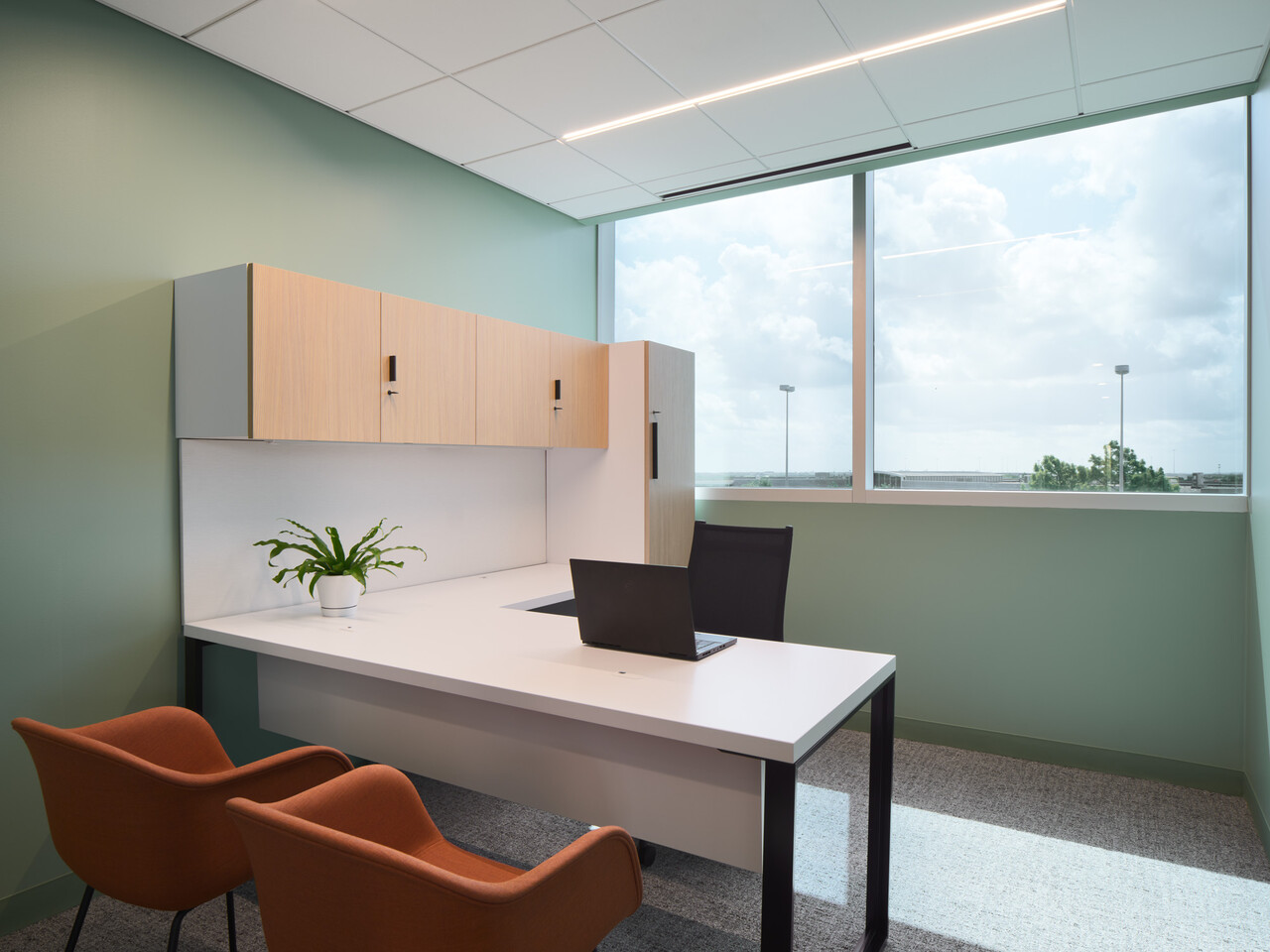 A modern office with a large window, white desk, black chair, two orange guest chairs, a laptop, a potted plant, and light wood cabinets against pale green walls and gray carpet.