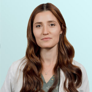 A woman with long brown hair wearing a white coat and a green blouse stands against a light blue background, looking calmly at the camera.