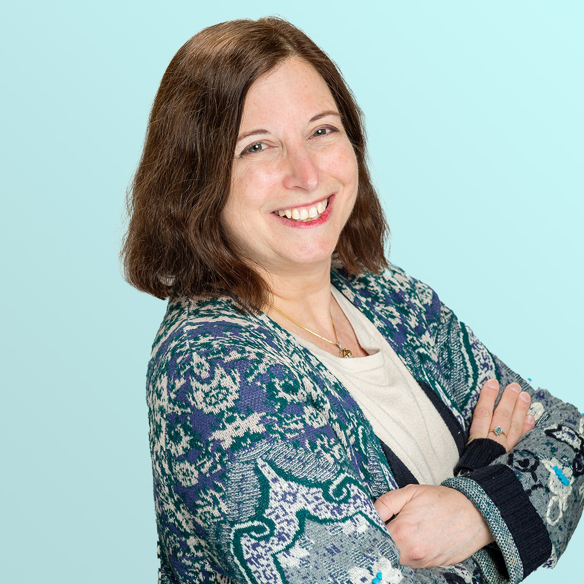 A woman with shoulder-length brown hair smiles with arms crossed, wearing a patterned blue and green sweater over a light top, against a light blue background.