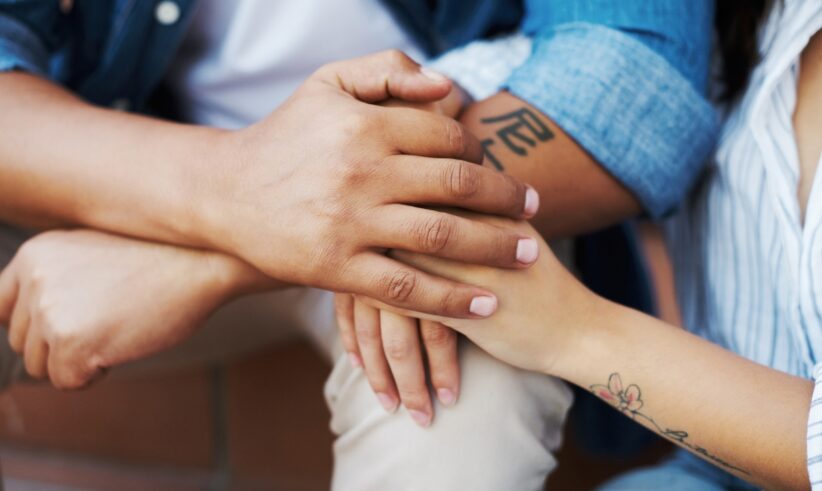 Close-up of two people sitting closely, gently holding hands in a comforting gesture. One person has a tattoo on their forearm and the other has a floral tattoo on their wrist. Both are wearing denim shirts.