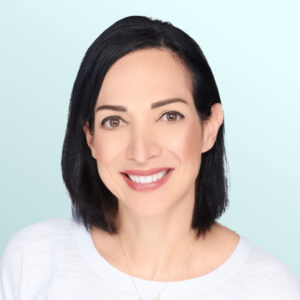 A woman with straight dark hair and light skin smiles at the camera. She is wearing a white shirt and a delicate gold necklace, and the background is a soft, light blue gradient.
