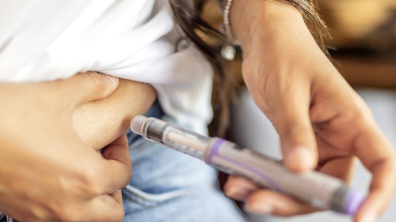 A person in jeans and a white shirt injects medication into their abdomen using an insulin pen. Their hand pinches the skin, and several bracelets are visible on their wrist.