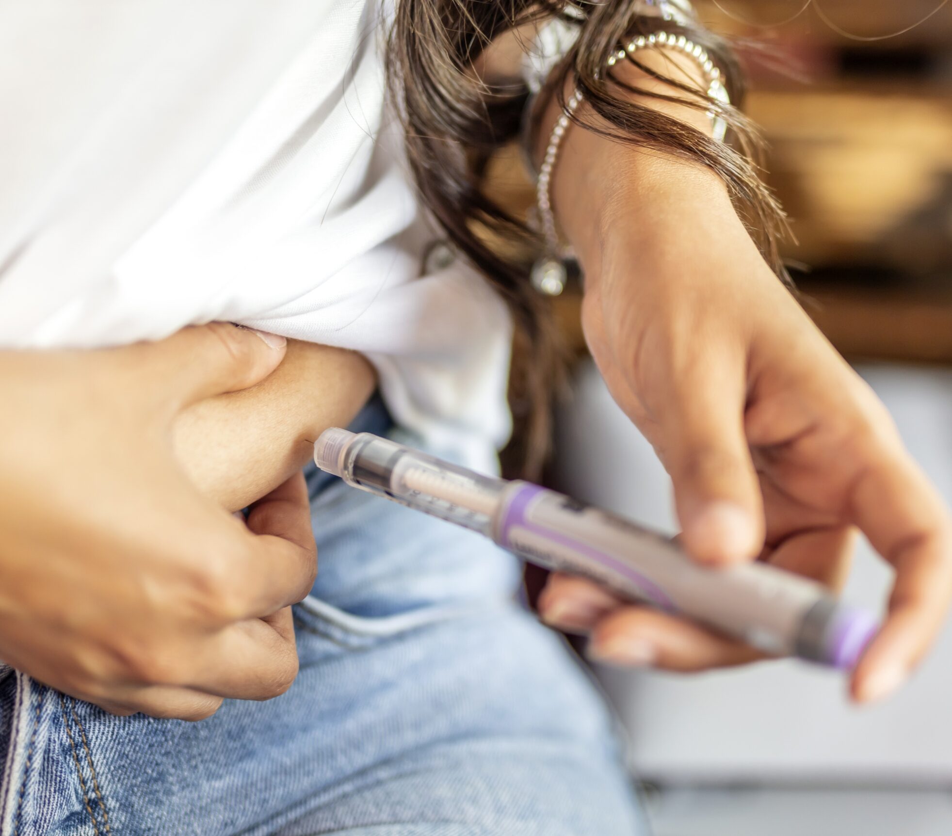 A person in jeans and a white shirt injects medication into their abdomen using an insulin pen. Their hand pinches the skin, and several bracelets are visible on their wrist.