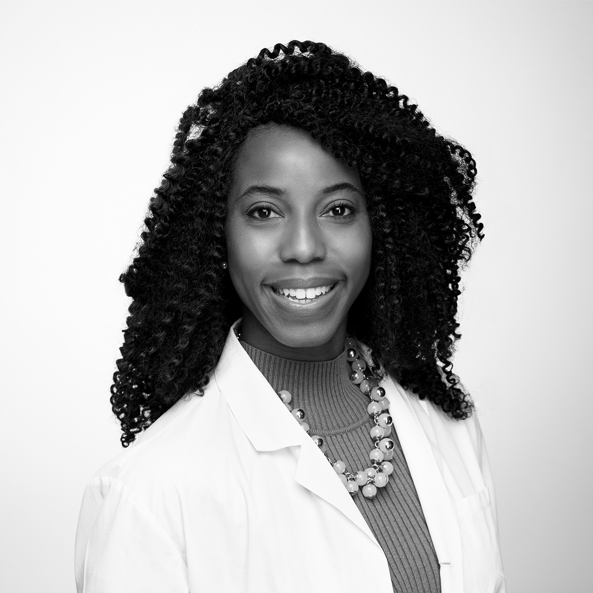 Black and white portrait of a woman smiling, wearing a white lab coat, a beaded necklace, and a ribbed turtleneck sweater. She has curly hair and is posed against a plain background.