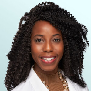 A smiling woman with curly hair wearing a white lab coat, a brown turtleneck, and a pearl necklace, posing in front of a light blue background.