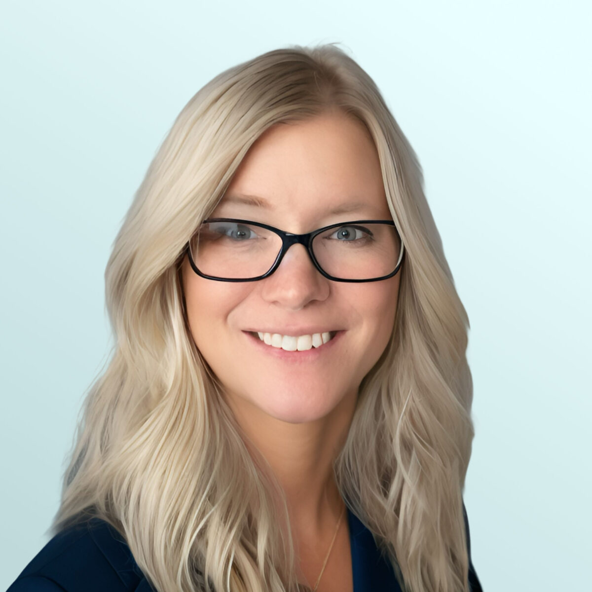A woman with long, wavy blonde hair and glasses smiles at the camera. She is wearing a dark blazer and a necklace, with a light blue background.