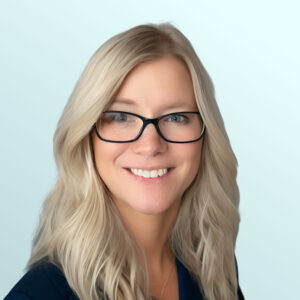 A woman with long, wavy blonde hair and glasses smiles at the camera. She is wearing a dark blazer and a necklace, with a light blue background.