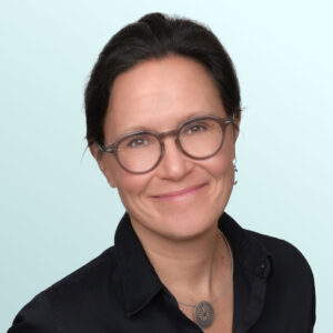 A woman with dark hair pulled back, wearing glasses, a black collared shirt, and a round pendant necklace, smiles at the camera against a light blue background.