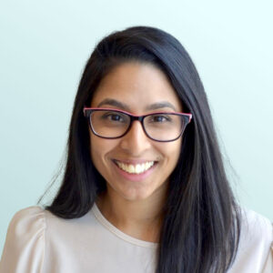 A woman with long dark hair and glasses smiles at the camera. She is wearing a light-colored blouse and is posed in front of a plain light blue background.