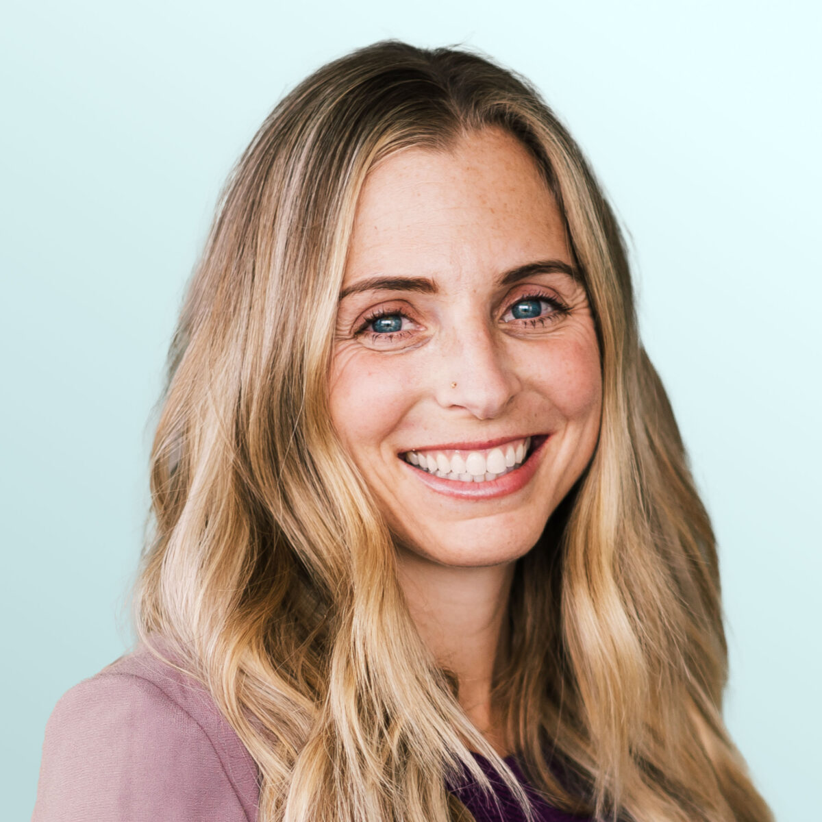 A woman with long, wavy blonde hair smiles at the camera. She is wearing a mauve jacket over a purple top and stands in front of a light blue background.