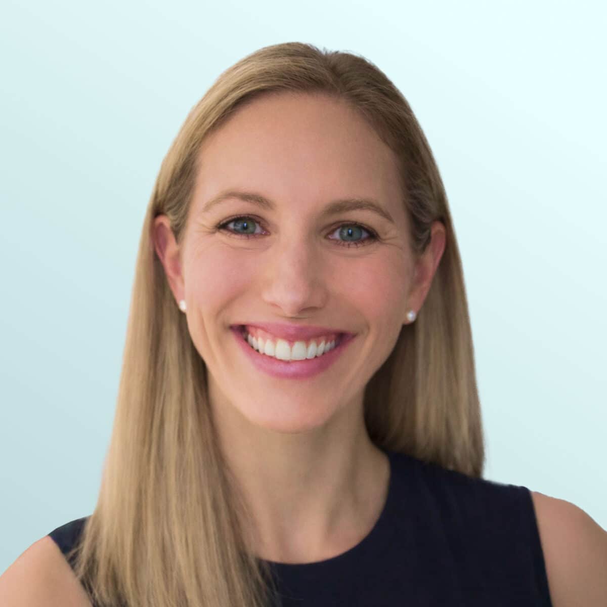 A woman with straight, blonde hair smiles at the camera. She is wearing a sleeveless dark top and pearl earrings, standing against a light blue background.