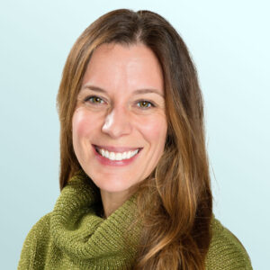 A woman with long brown hair, wearing a green turtleneck sweater, smiling against a light blue background.