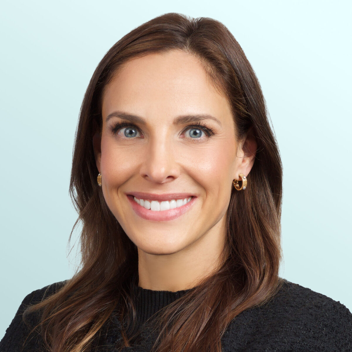 A woman with long brown hair wearing a black textured top and gold hoop earrings smiles at the camera against a light blue background.