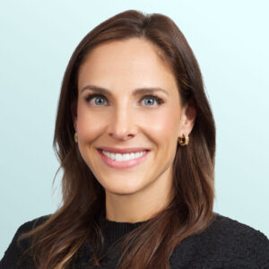A woman with long brown hair wearing a black textured top and gold hoop earrings smiles at the camera against a light blue background.