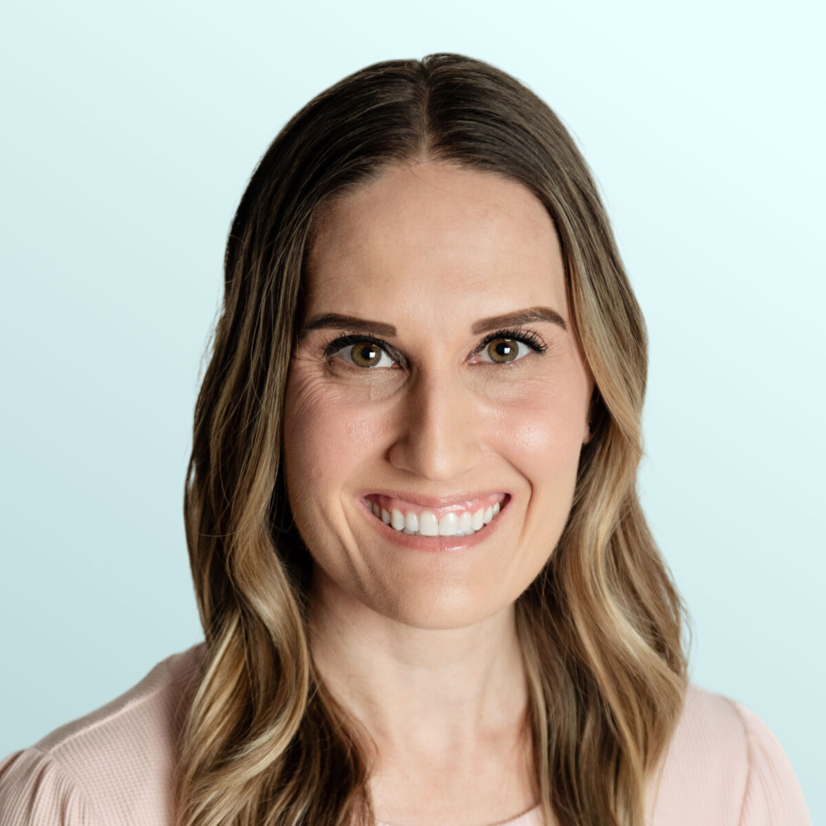 A woman with long, wavy brown hair and light highlights smiles at the camera. She is wearing a light pink top and is posed in front of a pale blue background.