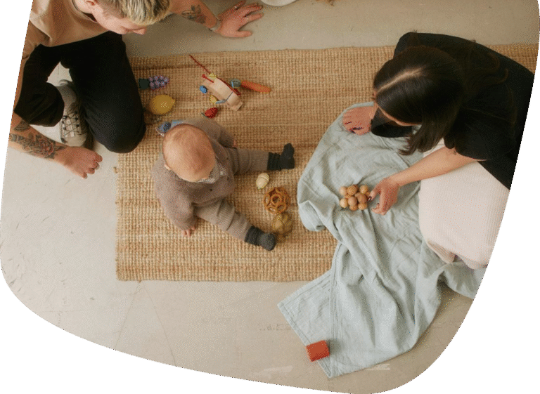 A baby sits on a woven rug playing with wooden toys, while two adults kneel nearby, watching and interacting with the child. A blue blanket and more toys are spread out beside them.